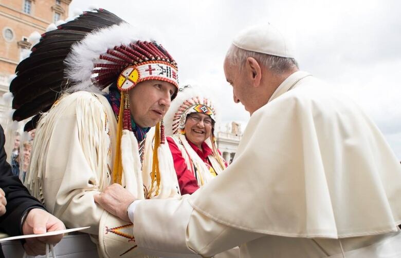 A man in a First Nations headdress shakes hands with a man in white papal robes.