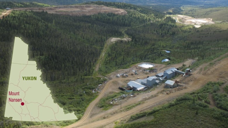 A group of mining buildings along a resource road in the foreground. In the background, rolling, green hills. As well, the open pit of the mine.