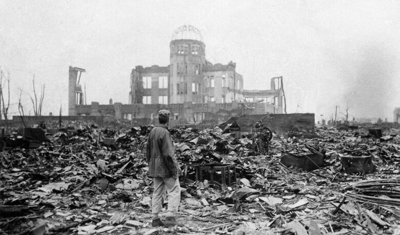 A black and white photograph of a man standing in front of the shell of a destroyed building, with the damaged frame of a dome on top. 