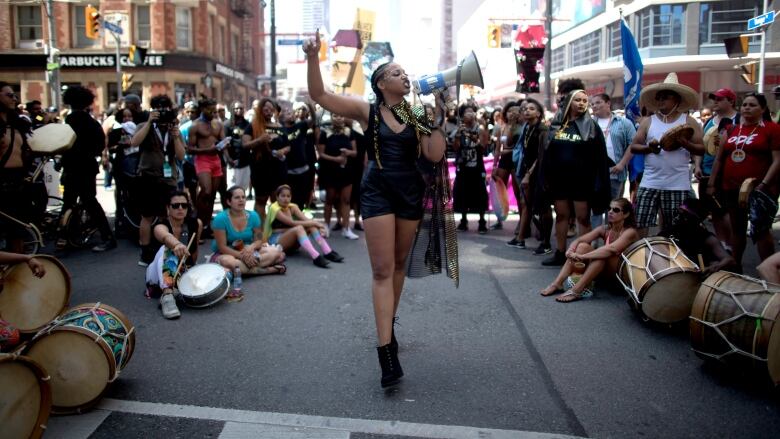 A person speaking through a megaphone on a road is in the centre of a circle of people who are listening to them.