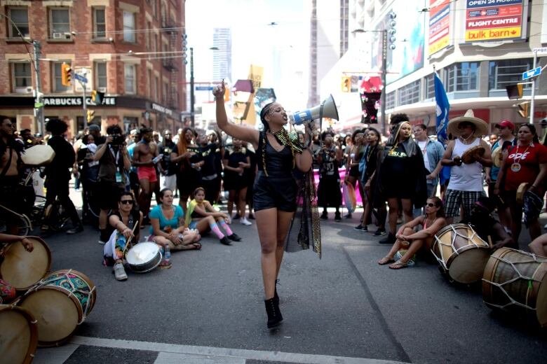 A person speaking through a megaphone on a road is in the centre of a circle of people who are listening to them.