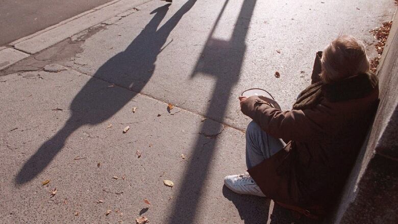 A man crouches down on a city sidewalk, holding  a hat in his hand, while the shadow of a person walking past him is seen.