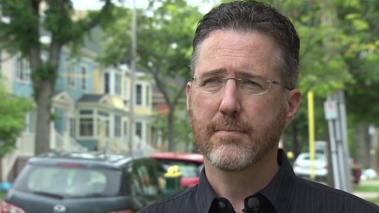 A man in a black shirt and glasses stands on a street.