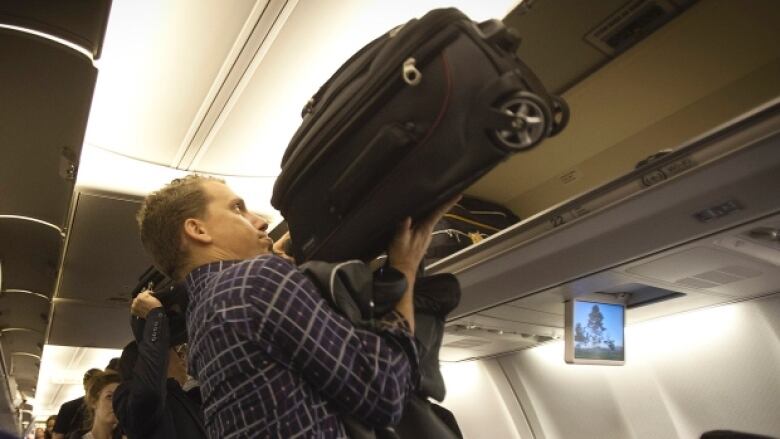 A man lifts a small black suitcase into the overhead bin in an airplane cabin.