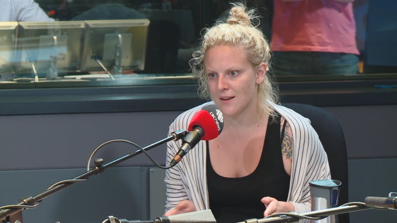 The photo of Mandi Gray was taken in 2016. She is in a CBC News studio with a radio mic in front of her. She's wearing a black tank top and stripped shirt.