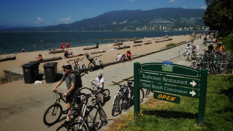 A group of bicyclists pass a beach, where people are sitting on logs.