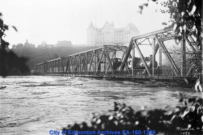 Black-and-white photo of a railway bridge with fast-flowing water right beneath it. 