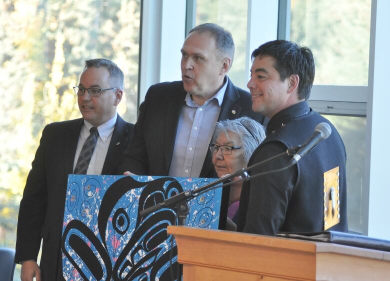 Three men and an elderly woman pose beside a podium, holding a piece of Indigenous artwork.