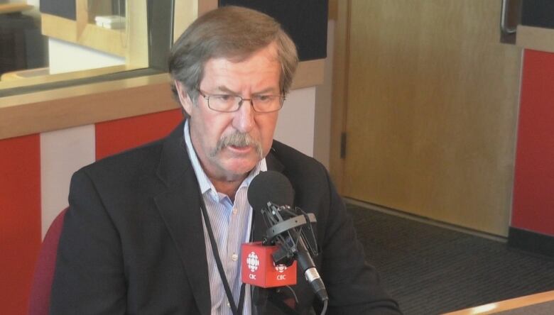 A man with light brown hair, a moustache and glasses talks into a microphone in a radio studio.