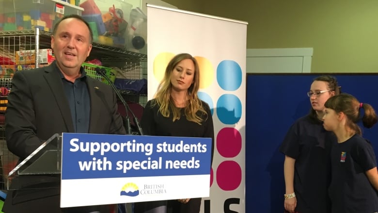 A man at a podium with a kid sitting underneath it and a sign across the front of the podium that says, Supporting students with special needs.