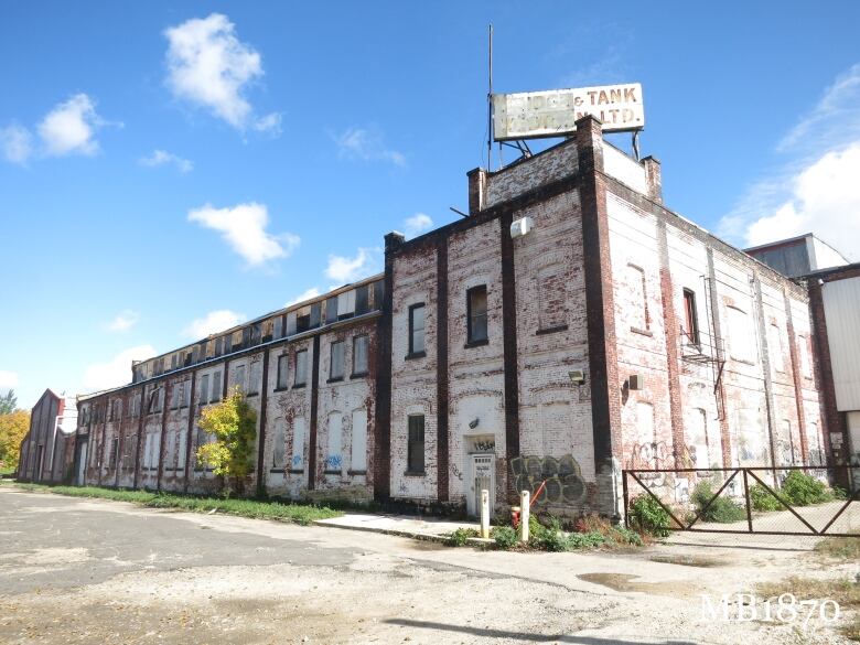 A large brick building with peeling paint, boarded windows and a half-unreadable sign on its roof sits beside a roadway.