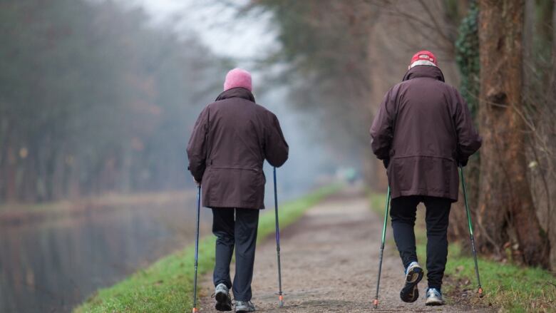 Two seniors use walking sticks to walk down a forest path.