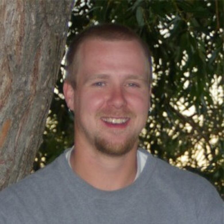 A man in grey stands in front of a tree.