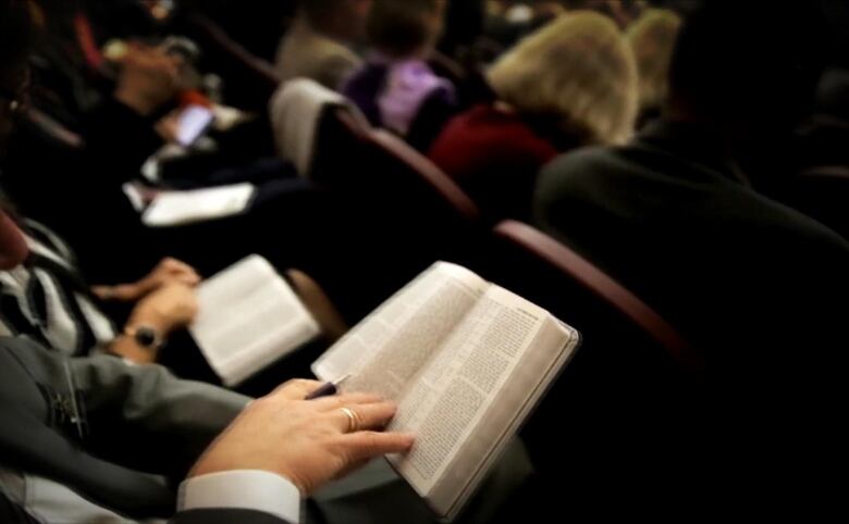 A scene from a Jehovah's Witness congregation shows people sitting in rows, reading from Bibles.