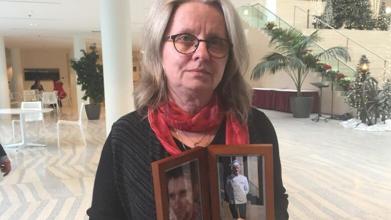 An older woman wearing glasses has a neautral expression while holding a picture frame of her son.