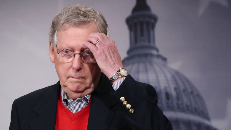 An older man in a suit gestures while standing in front of an image of the U.S. Capitol dome. 