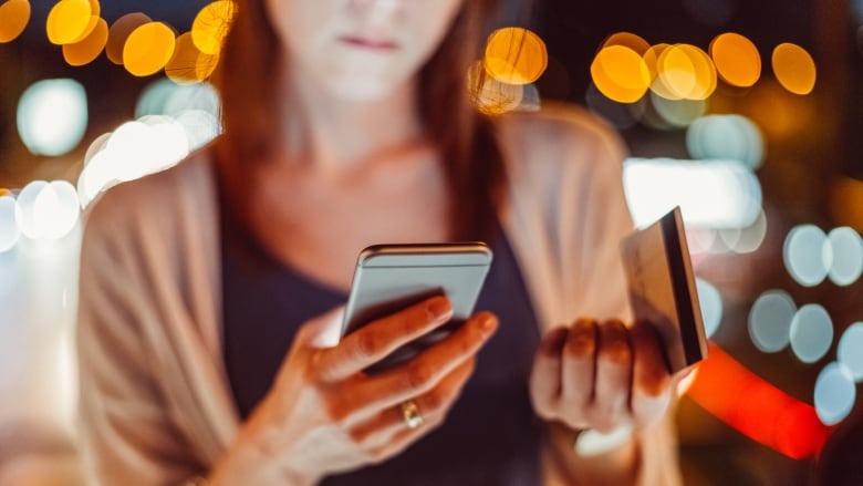 A woman looks at her cell phone while holding a credit or bank card in her hand.