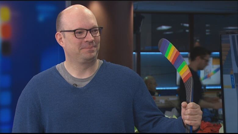 A man in a blue sweater holds a hockey stick wrapped in rainbow-coloured tape.