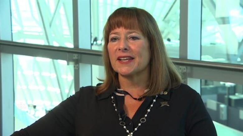 A woman with bangs talks in front of some windows. 