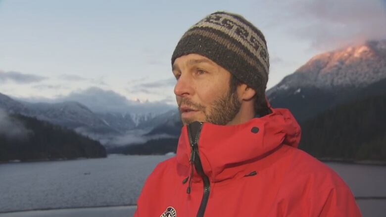 A man in red jacket and a beanie interviewing for camera with snow-capped mountain range and river on the background. 