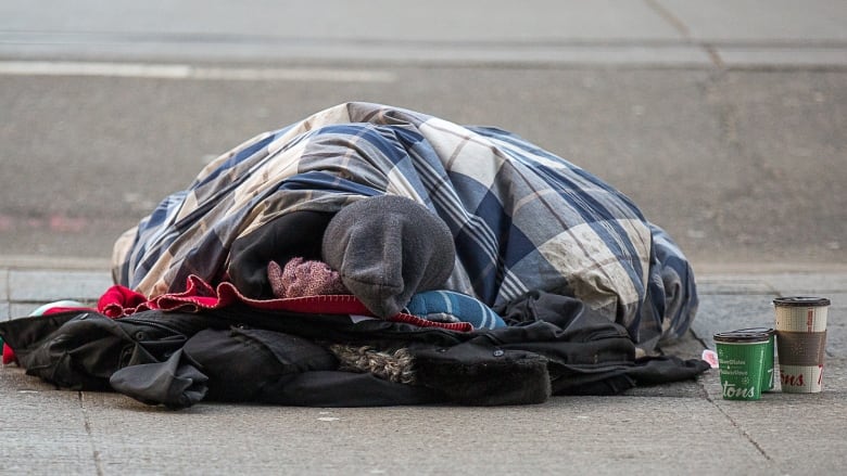 A homeless man sleeps in a sleeping bag on the sidewalk.