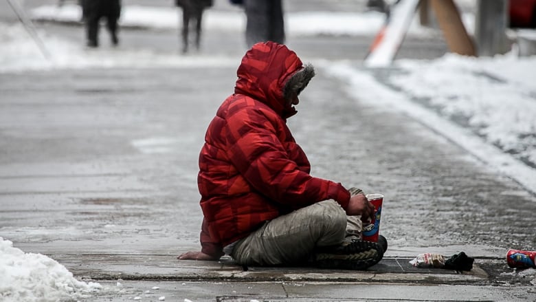 A man in a red winter coat and no gloves sits on a ice covered sidewalk.