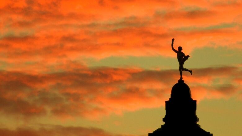 In front of a sunrise of casting orange and pink hues on clouds is a silhouette of the Golden Boy atop the Manitoba Legislature.