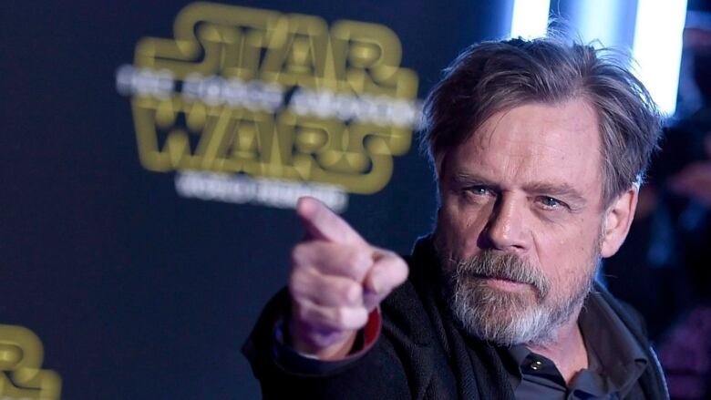 A man with a beard points just off camera as he stands in front of a backdrop with the Star Wars logo.