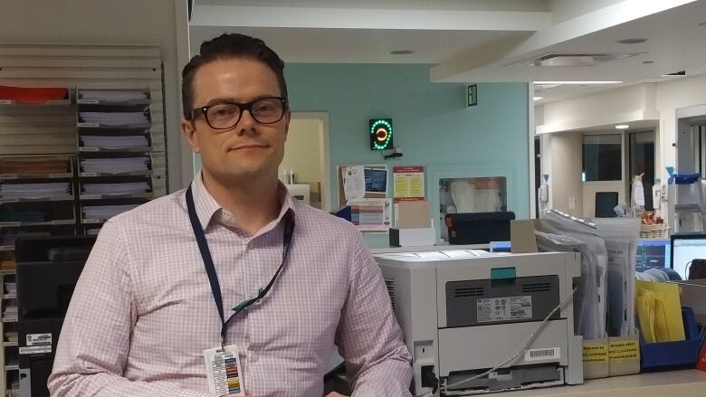 A man with short brown hair wearing a pink dress shirt and glasses stands near a desk at a hospital room. He's wearing a lanyad with hospital identifications.