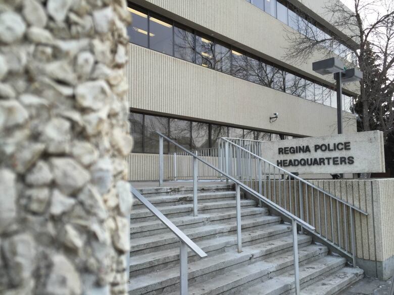 A sign in front of a stone building reads, 'Regina Police Headquarters.' There are stairs with metal rails in front of the sign.