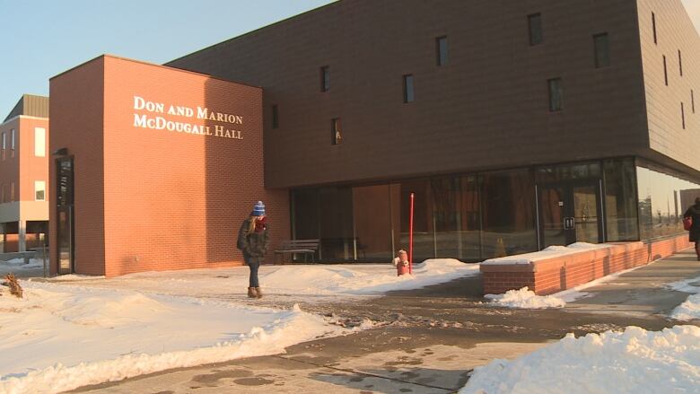 A woman in a winter coat and hat walks in front of a large brick building labelled Don and Marion McDougall Hall. 