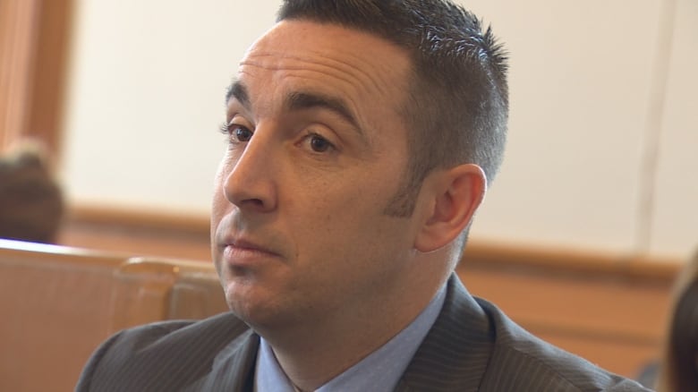 Close up of man's face, he's wearing a dark suit and light blue dress shirt. There's wood paneling behind him.