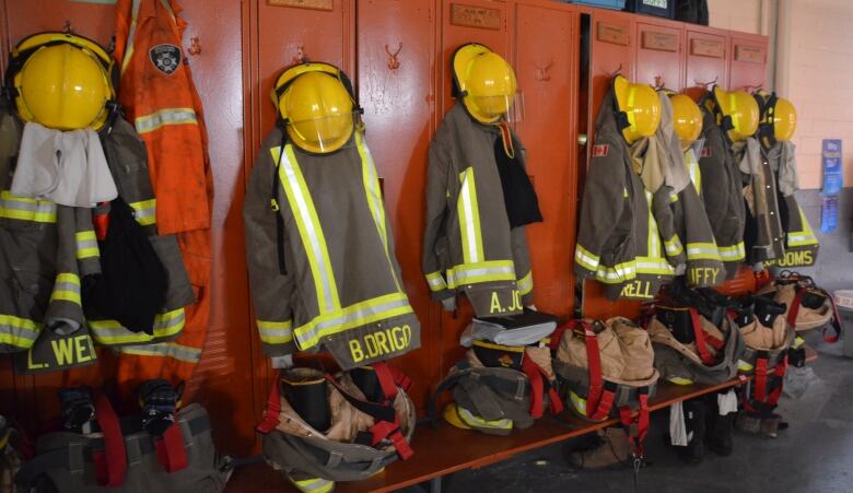 Firefighter helmets, coats and boats hang in a row on some orange lockers in a fire hall