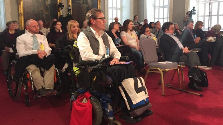 A group of people seated in a large room with red carpet, some in chairs and some in wheelchairs. 