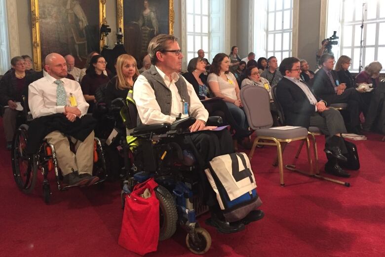 A group of people seated in a large room with red carpet, some in chairs and some in wheelchairs. 