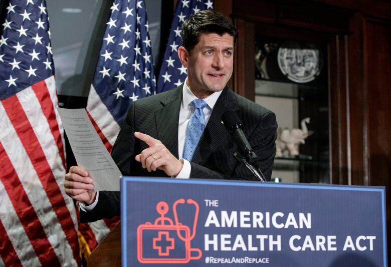 A man wearing a suit gestures toward a piece of paper he's holding in his right hand while speaking at a podium. In the background are three red, white and blue American flags. A placard on the podium reads: The American Health Care Act. # Repeal and Replace.