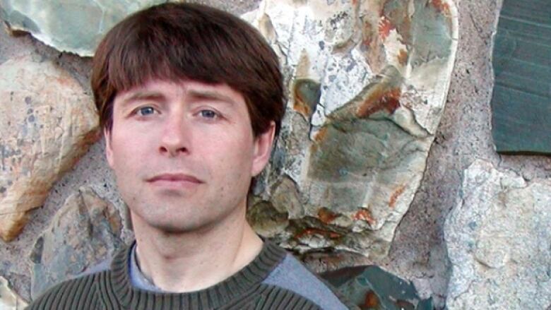 A close-up photo of a man with a bowl cut standing in front of a stone wall.