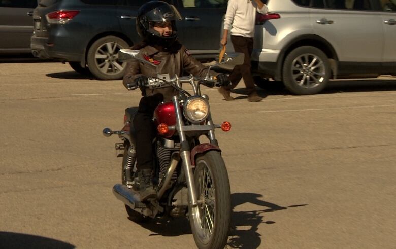 A woman wearing a helmet and leather jacket rides a motorcycle through a parking lot.