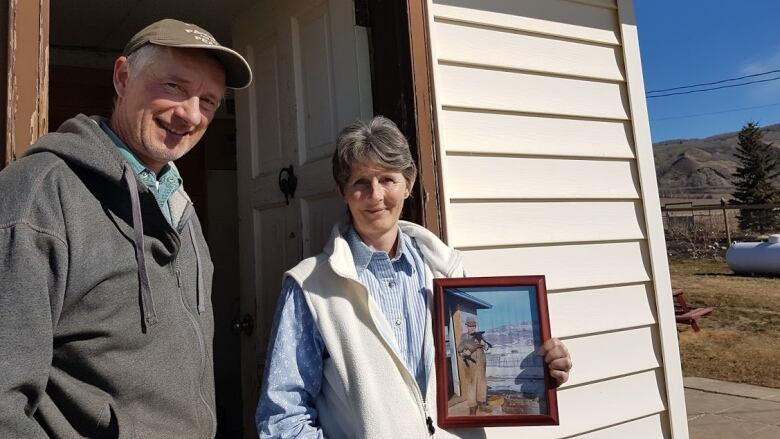 Two people stand at the door of their house, with one of them holding an old picture.