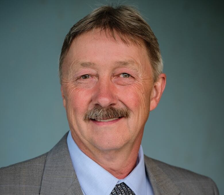 A portrait of a middle-aged man with a mustache, wearing a grey suit with a medal on the lapel, a light blue shirt and dark blue patterned tie.