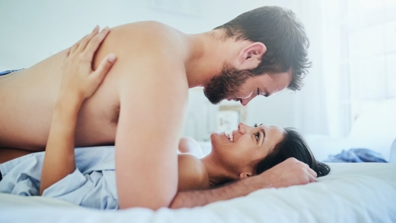 A man lays on top of a woman on a white bed. They smile at each other.