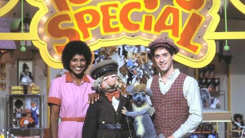 A still from a TV show featuring a smiling Black woman in a pink jumper next to a man in a hat, and two puppets. 