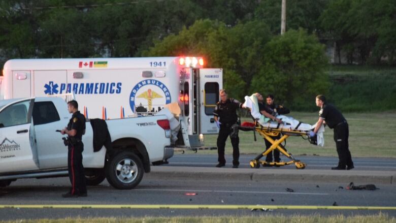 Two paramedics load a person on a gurney into the back of an ambulance at the scene of vehicle crash on a street.