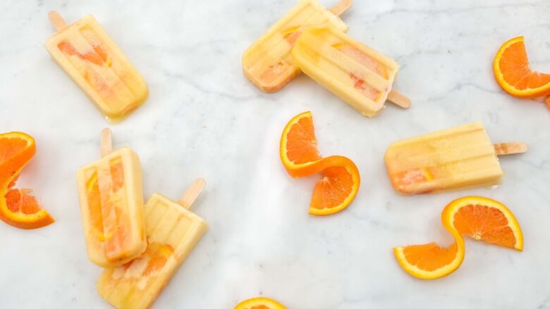 overhead shot of 6 creamy, orange popsicles and orange slices laying on a white marble surface. 