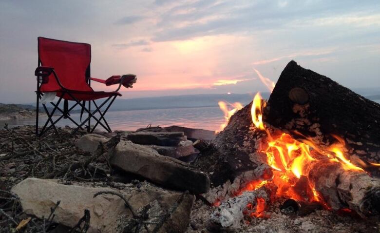 An empty red camping chair sits in front of a burning campfire.