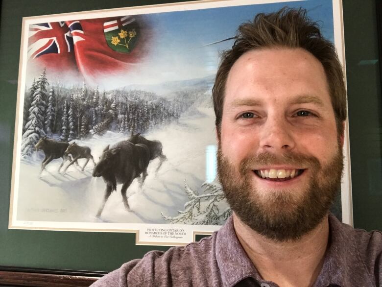 A bearded man smiles as he stands in front of a painting of four moose charging through a snowy forest. 