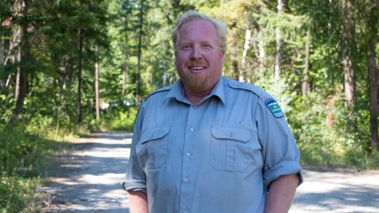 A man with a light red beard and blond hair in a blue open-collared shirt smiles at the camera framed by the forest behind him.