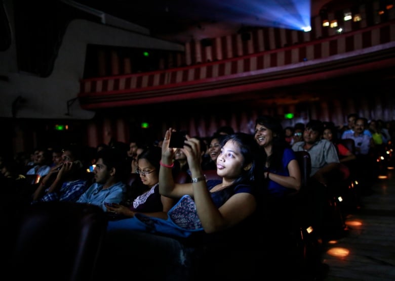 A girls face is illuminated by her phone screen as she takes a photo in a movie theatre. Behind her is a woman laughing while watching the screen. 