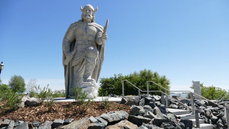 A statue of a 15 foot viking wearing a helmet and holding a staff towers over a small round park space against a blue sky. There are rocks and bushes surrounding him.  