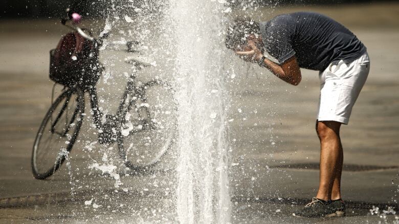 A man bends over with his hands on his face as he uses water from a fountain to cool down. His bicycle is nearby.